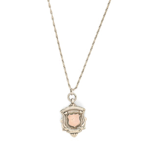 This shield charm is made of English sterling silver and features a rosy shield surrounded by a smooth ribbon border. Made in Birmingham, England in 1934, this piece bears the hallmarks for its date (L), silver material (lion), and location (anchor). Paired with one of our antique silver chains, this charm can be worn both as a pendant or in a cluster of charms. Front charm view, on chain