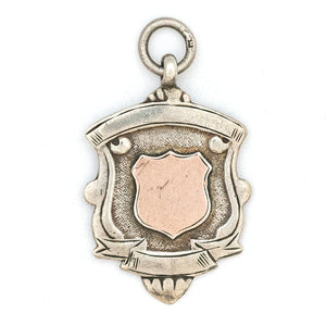 This shield charm is made of English sterling silver and features a rosy shield surrounded by a smooth ribbon border. Made in Birmingham, England in 1934, this piece bears the hallmarks for its date (L), silver material (lion), and location (anchor). Paired with one of our antique silver chains, this charm can be worn both as a pendant or in a cluster of charms. Front charm view