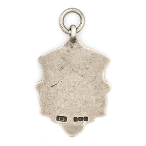 This shield charm is made of English sterling silver and features a rosy shield surrounded by a smooth ribbon border. Made in Birmingham, England in 1934, this piece bears the hallmarks for its date (L), silver material (lion), and location (anchor). Paired with one of our antique silver chains, this charm can be worn both as a pendant or in a cluster of charms. Back charm view, showing english hallmarks