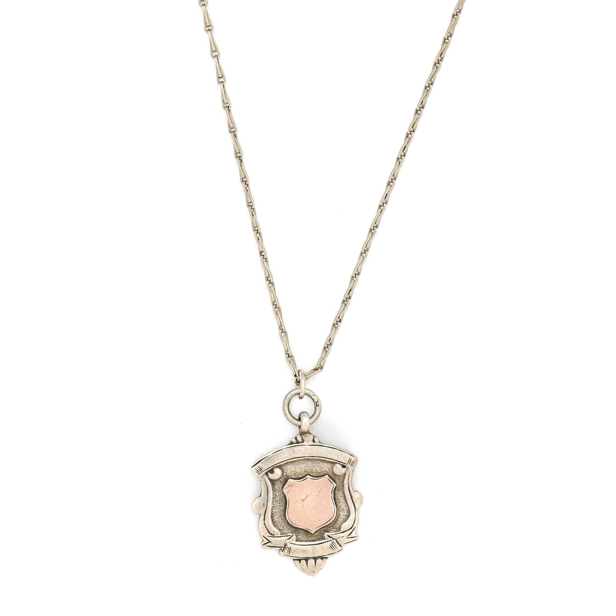 This shield charm is made of English sterling silver and features a rosy shield  surrounded by a smooth ribbon border.  Made in Birmingham, England in 1934, this piece bears the hallmarks for its date (L), silver material (lion), and location (anchor). Paired with one of our antique silver chains, this charm can be worn both as a pendant or in a cluster of charms.  Front charm view, on chain