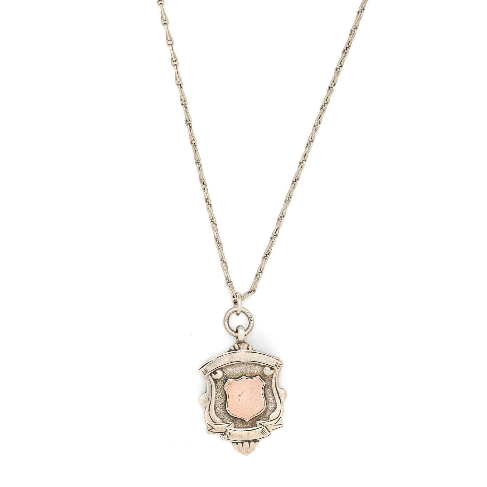 This shield charm is made of English sterling silver and features a rosy shield  surrounded by a smooth ribbon border.  Made in Birmingham, England in 1934, this piece bears the hallmarks for its date (L), silver material (lion), and location (anchor). Paired with one of our antique silver chains, this charm can be worn both as a pendant or in a cluster of charms.  Front charm view