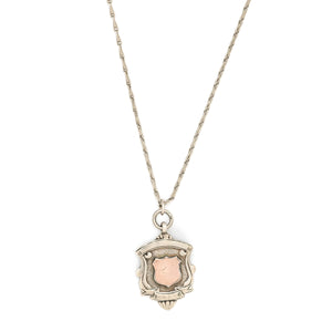 This shield charm is made of English sterling silver and features a rosy shield  surrounded by a smooth ribbon border.  Made in Birmingham, England in 1934, this piece bears the hallmarks for its date (L), silver material (lion), and location (anchor). Paired with one of our antique silver chains, this charm can be worn both as a pendant or in a cluster of charms.  Front charm view, on chain
