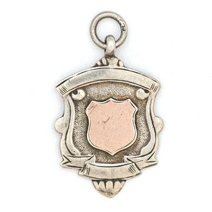 This shield charm is made of English sterling silver and features a rosy shield  surrounded by a smooth ribbon border.  Made in Birmingham, England in 1934, this piece bears the hallmarks for its date (L), silver material (lion), and location (anchor). Paired with one of our antique silver chains, this charm can be worn both as a pendant or in a cluster of charms.  Front charm view